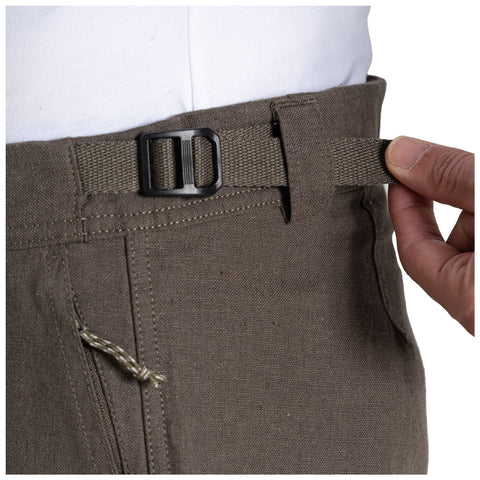 A person adjusts a brown fabric belt on their trousers using their right hand while wearing a light-colored shirt in a neutral outdoor or indoor setting