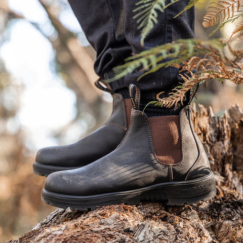 Brown leather boots are positioned on a textured wooden surface surrounded by greenery with light filtering through the trees creating a natural outdoor setting.