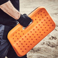 An orange padded kneeling pad is being held by a person wearing a black glove. The hand is gripping the pad as they stand on a rustic, sandy surface.