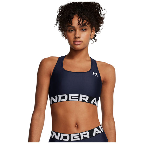 A young woman with curly hair is standing confidently wearing a navy blue sports bra and matching bottoms featuring a prominent brand logo. The background is plain white.