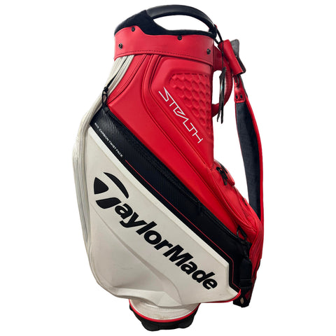 A golf bag designed for carrying clubs features a sleek design with a red and white color scheme and the logos of TaylorMade and Stealth. It includes a handle and a shoulder strap for easy transport.