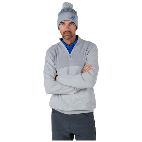 A man stands with arms crossed wearing a gray pullover and a beanie with a pom-pom. He appears focused and is in a neutral, indoor setting.