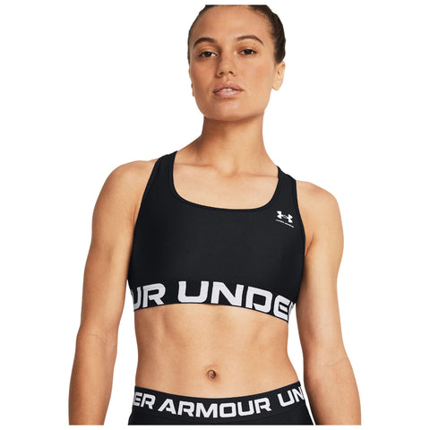 A woman poses confidently wearing a fitted black sports bra with a wide white band that displays the brand name Under Armour in large letters, set against a plain background.