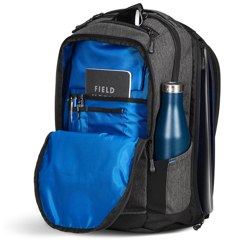 A gray backpack with a blue interior contains a notebook labeled FIELD NOTES pens and a water bottle in its side pocket while unzipped and open in a tidy context.