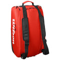 A red sports bag with black straps sits upright featuring a prominent white logo. It is designed for carrying sports gear in a travel or practice setting.