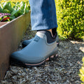 Gray gardening shoes are positioned on mulch next to a wooden planter filled with green plants while sunlight illuminates the scene and highlights surrounding greenery.