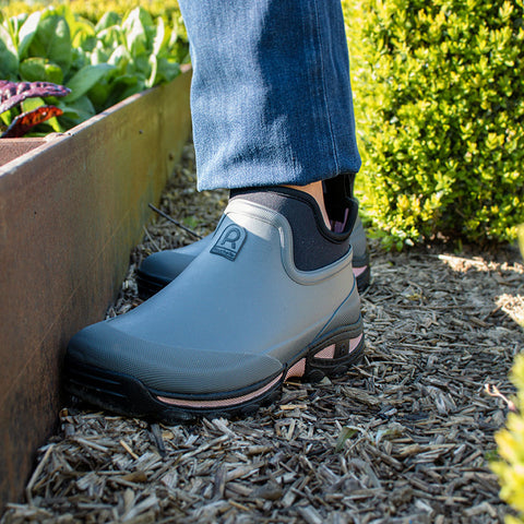 Gray gardening shoes are positioned on mulch next to a wooden planter filled with green plants while sunlight illuminates the scene and highlights surrounding greenery.