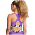 A woman is wearing a purple sports bra with a cutout back and a black and white band around her midsection in a neutral setting. Her curly hair is styled in a ponytail.