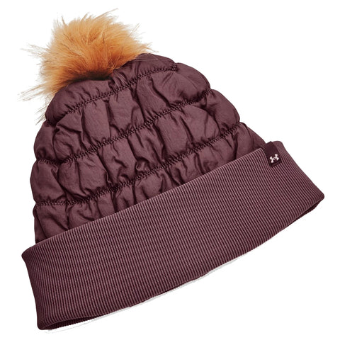 A puffy brown beanie with a fuzzy orange pom-pom sits tilted with a ribbed cuff at the bottom showcasing a logo highlighting its sporty design suitable for winter wear.