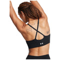 A woman in a black sports bra raises her arms, showcasing a world map tattoo on her back in a neutral background environment. The sports bra features the Under Armour logo.