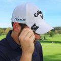 A man adjusts a white cap with Callaway branding while holding a small black item near the brim in a green outdoor golf setting with trees in the background.