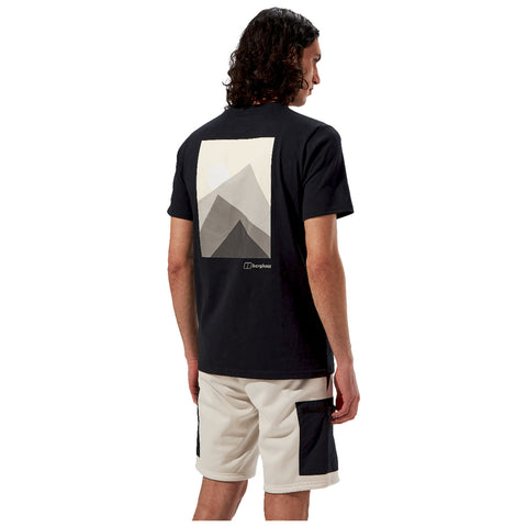 A person stands facing away wearing a black t-shirt featuring a geometric mountain design on the back paired with light shorts highlighting a casual, outdoorsy style.