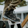 A black hiking boot rests on a metal ladder step showing signs of dirt and mud while a person in khaki pants is engaged in an outdoor task with a blurred background of a building.