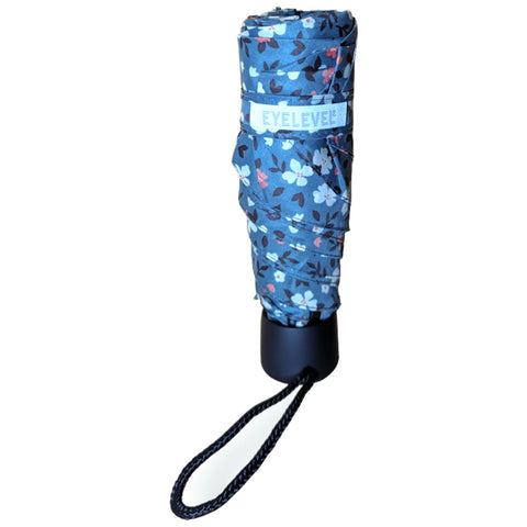 A compact floral-patterned umbrella is closed and resting upright with a black handle and a wrist strap attached displaying the brand label EYELEVEL around the middle section.