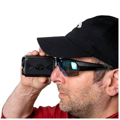 A man wearing sunglasses holds a black rangefinder up to his eye focused on a distant target in a bright or well-lit setting.