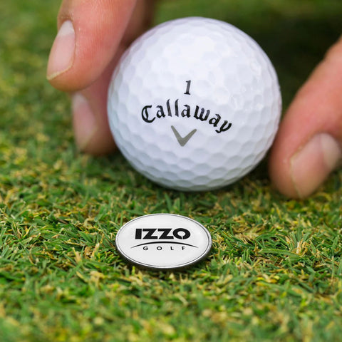 A white golf ball is being held above a small round marker labeled IZZO GOLF on green grass, indicating a setup for a golf shot.