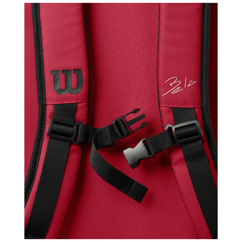 A red backpack is displayed with adjustable straps secured by a black buckle showing a logo and a signature on the surface highlighting a sporty design.