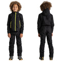 A child wearing a black outdoor jacket with yellow zippers and matching pants stands confidently. The backdrop is plain, focusing on the child’s stylish activewear and cheerful demeanor.