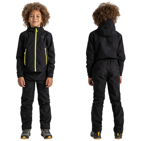 A child wearing a black outdoor jacket with yellow zippers and matching pants stands confidently. The backdrop is plain, focusing on the child’s stylish activewear and cheerful demeanor.