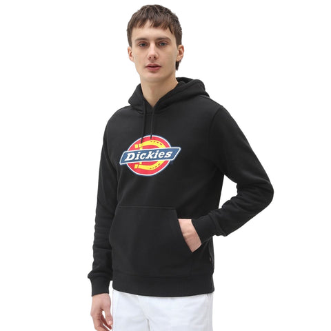 A young man stands wearing a black hoodie featuring a colorful logo at the center while casually placing one hand in the front pocket in a plain white background.