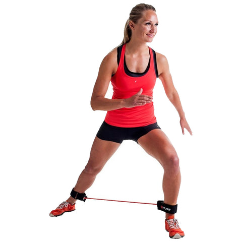 A woman in a red athletic tank top performs a lateral movement exercise while wearing resistance bands on her legs in a bright, minimalistic environment.