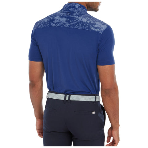 A person wearing a blue polo shirt with a patterned upper back is standing with their back facing the viewer, paired with dark trousers and a light-colored belt in a neutral setting.