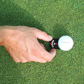 A hand holds a golf ball marker next to a white golf ball on a green lawn-like surface as the player prepares to mark the ball's position