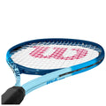 A blue tennis racket with a black handle rests in a slightly angled position showcasing a white string pattern and a prominent red logo on the strings indicating the brand.