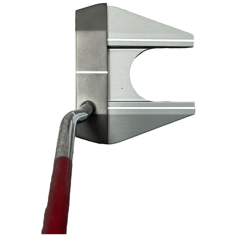 A metallic golf putter is positioned upright showcasing a wide club head with a slot and horizontal grooves while a curved handle extends backward providing grip.