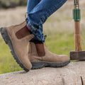 Brown boots stand on a log while a foot lifts slightly. Blue jeans fit snugly around the ankles. A hammer rests beside the log in a grassy outdoor setting.