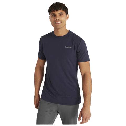 Man wearing a navy blue Calvin Klein t-shirt smiles while standing in a neutral background. His right hand is relaxed at his side while the left hand is slightly raised.