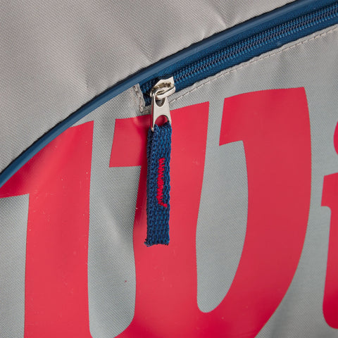 A zippered bag features a metal zipper pull attached to a blue fabric tab with red stitching on a surface displaying large red lettering against a gray background.