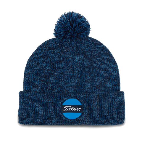 A navy blue knit beanie featuring a round blue and white patch with the word Titleist is displayed laid flat with a pom-pom on top against a plain background.