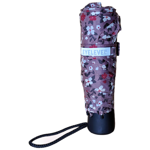 A closed umbrella with a floral patterned fabric wraps around the shaft while resting upright on its handle in a minimalistic setting, showing a black strap for carrying.