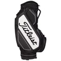 A black and white golf bag stands upright featuring a prominent logo with a textured design and multiple zippered pockets showcasing functionality in a sports setting.