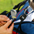 A hand holds a golf club cleaning brush while another hand grips a golf club near a golf bag on a green outdoor surface, suggesting preparation for use on the course.