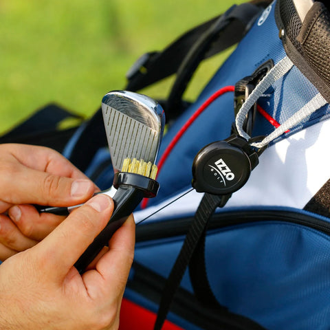 A hand holds a golf club cleaning brush while another hand grips a golf club near a golf bag on a green outdoor surface, suggesting preparation for use on the course.