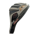 A golf club head features a shiny black and metallic finish with engraved details. It is angled upwards, ready for a swing on a golf course.