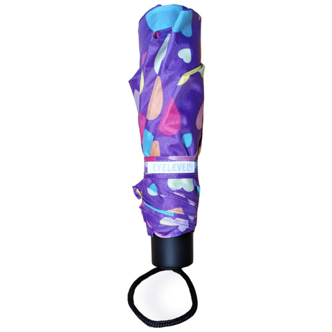 A colorful, closed umbrella features a purple fabric with various shapes in pink, blue, and yellow. It has a black handle and a wrist strap for easy carrying.