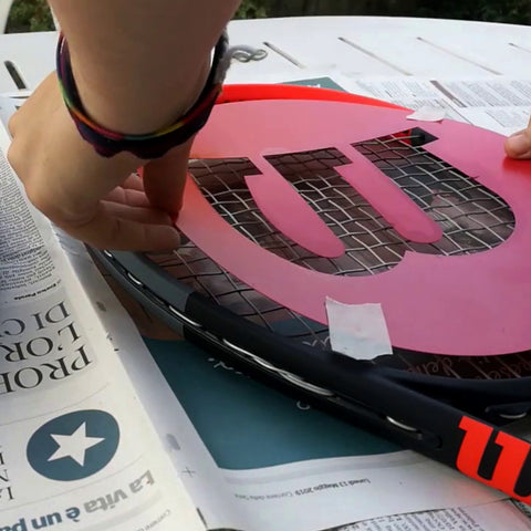 A hand places a pink stencil on a tennis racket lying on a table covered with newspaper while preparing to paint or mark the racket in an outdoor setting.