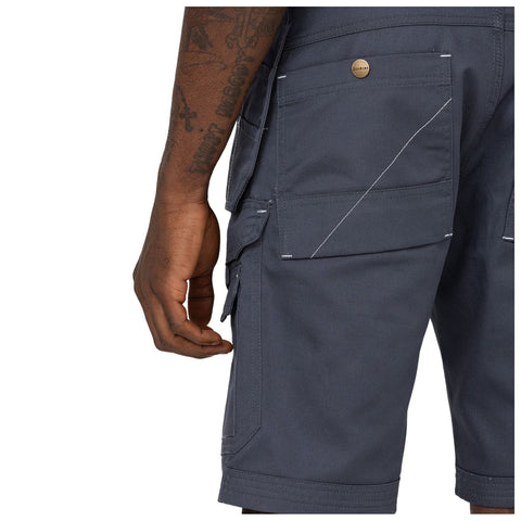 A hand is reaching towards the cargo shorts worn by a person showcasing side pockets and a button with a brand logo on a gray fabric background.