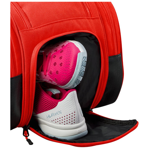 A red bag with a zippered compartment is opened revealing a pair of athletic shoes with pink and white detailing resting inside on a black fabric lining.