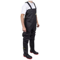 A person wearing black waterproof overalls stands upright with hands at their sides. They also wear black knee-high boots with red soles. The background is plain and white.