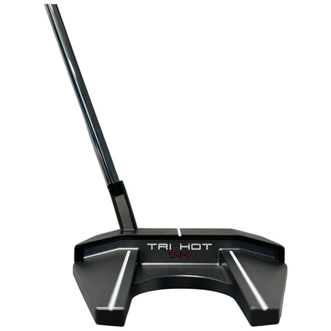 A black golf putter rests on a flat surface with a shiny metallic shaft angled upwards featuring a wide, flat head displaying the text TRI-HOT 5K and lined grooves for grip.