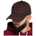 A brown baseball cap with a red logo is worn by a man with a beard who is dressed in a maroon hoodie and looking slightly downwards in a neutral setting.