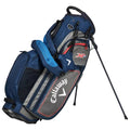 A blue and gray golf bag is standing upright with a retractable stand extended. It features multiple pockets and a padded shoulder strap for carrying golf clubs and accessories.