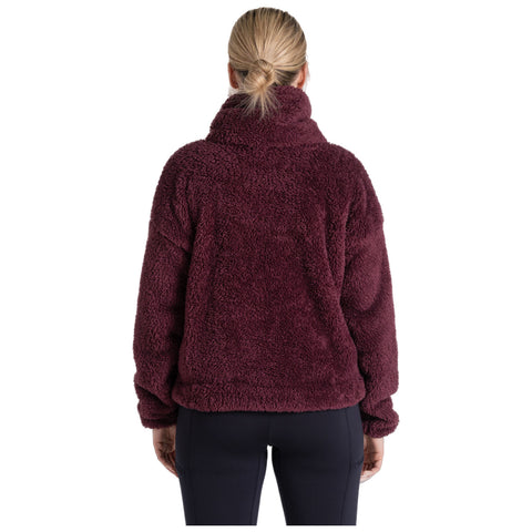 A woman is standing with her back facing the viewer, wearing a plush, deep burgundy pullover. She has her hair in a low ponytail and is in a neutral setting.