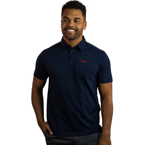 A man is smiling while standing with one hand on his hip he wears a dark blue polo shirt with a small red logo on the chest and has short curly hair against a plain white background.