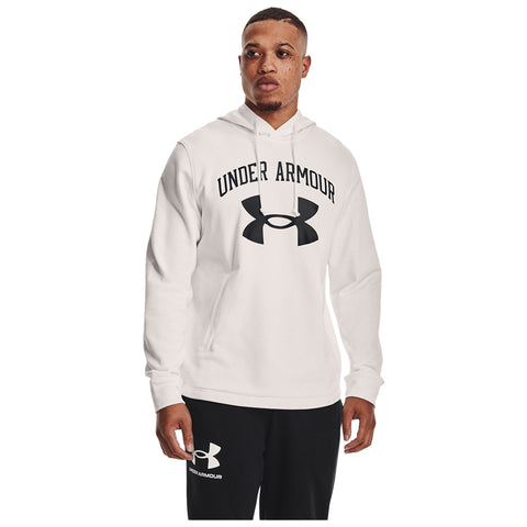 A man stands wearing a white hoodie with "UNDER ARMOUR" printed in black across the chest and black sweatpants with a logo near the ankle in a plain background.