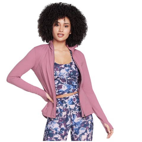 A woman with curly hair poses confidently while wearing a pink zip-up jacket over a colorful athletic outfit in a bright, simple backdrop that emphasizes her activewear style.